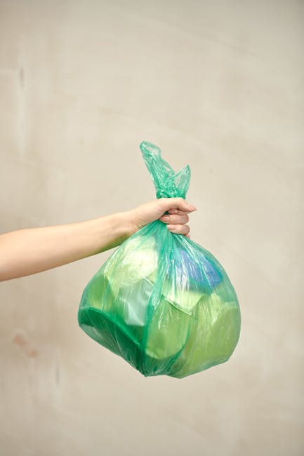 A person's hand and forearm are visible holding a green and blue recycling bag made of thin, translucent plastic material, tied at the top with a knot. The bag appears to be filled with lightweight waste, illustrating typical rubbish for collection or disposal. The background is plain and neutral, possibly a beige wall, with soft lighting that minimizes shadows. This image emphasizes the handling of waste in a manner suitable for private rubbish disposal or on-site clearance, potentially related to alternative waste management solutions offered by services such as wasteclearancekingstonuponthames.co.uk. The scene reflects the process of managing and preparing household or small-scale waste for removal, aligning with the context of rubbish removal services in Kingston upon Thames. The focus is on the bag's material, color, and the act of holding it, with no additional objects or environmental details shown, maintaining a professional and factual tone relevant to waste collection practices.