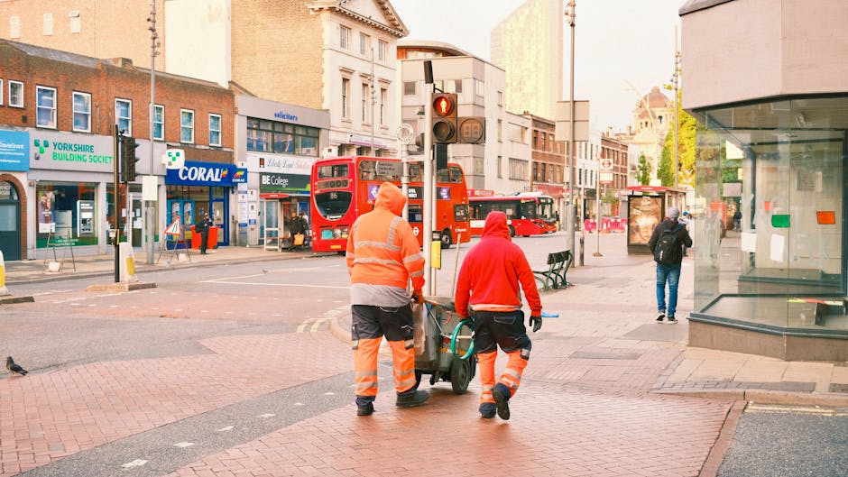 Two waste removal workers dressed in high-visibility orange and black uniforms are walking on a city sidewalk, pushing a wheeled trash bin towards the curb for collection. The worker on the left is wearing a jacket with reflective strips and dark trousers, while the worker on the right is wearing a red hoodie and matching high-visibility trousers. In the background, there are red double-decker buses stopped at a traffic light, along with various commercial buildings, including a pharmacy, a building society, and a college sign. Other pedestrians are visible, including one person in dark clothing walking away from the camera near a bus stop shelter. The scene is set during daylight hours with soft, natural lighting illuminating the urban environment, which appears organized and orderly, indicating an area where private waste collection services may operate as an alternative to civic rubbish collection. The overall setting highlights a typical busy urban street scene focused on waste management activities, natural environmental context, and city infrastructure.