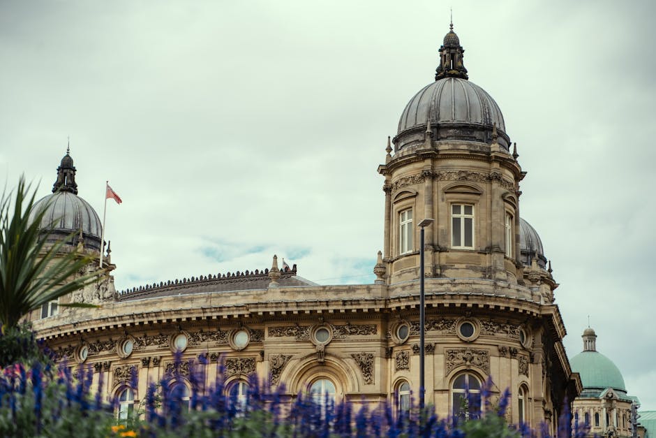A close-up view of a historic stone building with ornate architectural details, including large domed rooftops topped with smaller spires, set against a cloudy sky. The structure features classical design elements such as arched windows, decorative cornices, and intricate carvings along the facade, constructed from beige and light-colored stone with a weathered finish. In the foreground, blurred purple flowers and green foliage partially obscure the lower part of the building, adding natural contrast to the historic architecture. A slim street lamp stands on the pavement nearby, with no visible vehicles or people present in the scene. The environment appears to be part of an urban area, possibly near a park or public square, with subtle hints of greenery surrounding the building. This scene emphasizes the durability and detailed craftsmanship of the historic architecture, suitable for context around vintage and private waste removal services that work alongside such cityscapes, subtly highlighting an environment where on-site or alternative disposal methods may be relevant.