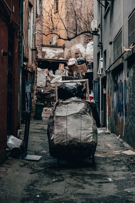 A large pile of black rubbish bags piled against a modern tiled wall in an outdoor urban setting. The bags appear to be filled with mixed waste, some with plastic bottles and other small debris visible on and around them. The bags are loosely arranged, with some leaning against each other and others resting directly on the ground. Behind the pile, the wall has a graffiti spray-painted message in red reading 'XEND'. To the left, a section of a metallic cylindrical structure is partially visible, and the background shows cloudy sky and distant buildings, indicating an area experiencing unmanaged waste accumulation. The scene suggests an incident of illegal dumping or overflow of waste collection, relevant to private rubbish removal services that handle excess or blocked access waste collection in Kingston and similar urban areas.