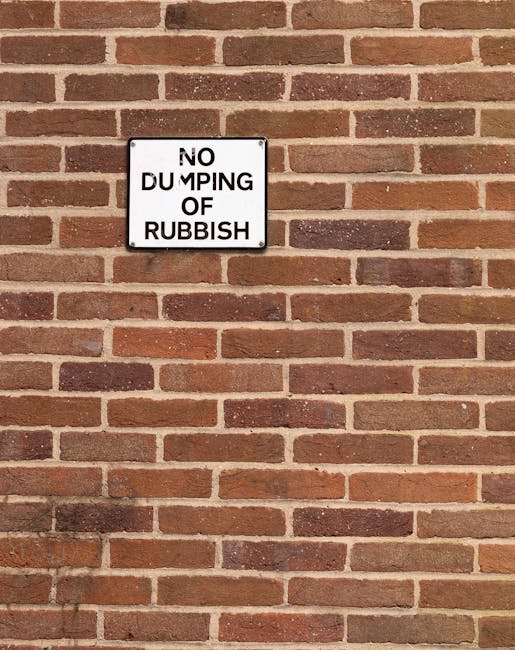 A rectangular metal sign mounted on a red brick wall displaying the words 'NO DUMPING OF RUBBISH' in bold black capital letters with some letters slightly misaligned. The brick wall features a traditional running bond pattern with reddish-brown bricks and dark mortar joints. The lighting appears natural, casting subtle shadows below the sign. The scene suggests a controlled environment where waste removal and disposal are managed, possibly indicating restrictions on private rubbish dumping to support cleaner, organized waste management practices. The sign’s placement on the exterior wall aligns with independent waste handling considerations, subtly connecting to rubbish removal services such as those offered by waste clearance companies in Kingston upon Thames to support community and environmental standards.