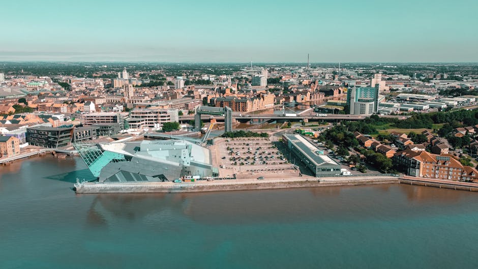 An aerial view of a cityscape showing a large modern building with angular, metallic grey and teal facades situated on the riverbank, with reflections visible in the water. Adjacent to the building, there is a spacious open area with a parking lot partly filled with cars, and an elongated, low-rise structure with dark windows. Surrounding the waterfront are mixed-use buildings, including several red-brick residential developments and commercial structures. The background reveals a densely built urban area with numerous mid-rise and high-rise office and apartment buildings, as well as bridges spanning the river, connecting different parts of the city. The scene is captured in daylight under clear or lightly overcast skies, highlighting the varied textures and colors of the city’s architecture, set within a practice of urban development that could relate to alternative or private waste management facilities situated within a busy city environment for efficient rubbish removal services in Kingston upon Thames.