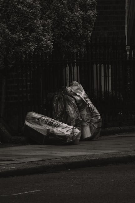 Three large black garbage bags filled with waste are placed on the pavement outside, leaning against a black metal fence that encloses a garden area with dense foliage. The bags appear to be made of thick plastic and are slightly crumpled, with visible fold lines and some tearing or damage at the top. The surrounding environment includes a section of road in the foreground with a curb separating the pavement from the street. Behind the fence, there are trees or bushes with dark, leafy textures, partially obscured by shadows. The scene is lit with soft natural light, suggesting an overcast day or shaded area. The image exemplifies a typical setup for private rubbish disposal outside a residential property, aligning with non-collection waste handling practices often performed by waste clearance services such as those offered by wasteclearancekingstonuponthames.co.uk, which facilitate alternative waste management solutions outside public rubbish collection rules.