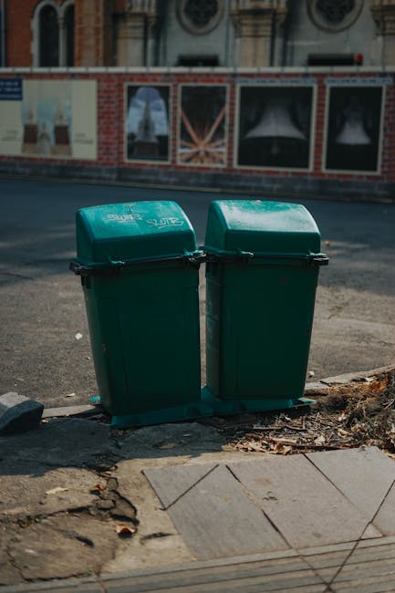 Two green plastic waste bins with hinged lids are positioned side by side on a paved pavement at the edge of a street, with their backs facing the road. The bins are made of textured, durable plastic and feature black clasp mechanisms securing the lids. The pavement surface beneath the bins shows signs of wear, with a few scattered dried leaves and small debris nearby. In the background, there is a brick and painted wall with partially visible posters, advertising a building or event, and above it, a row of windows with decorative frames. The street appears quiet, with a slightly blurry view of the roadway and a parked vehicle visible in the distance. The scene suggests a typical urban environment where private rubbish collection or on-site waste management may be employed, supported by services like wasteclearancekingstonuponthames.co.uk, which handles waste disposal outside of local authority recyclables. Lighting is natural daylight, casting subtle shadows, and the overall atmosphere remains neutral and clean, emphasizing the functional role of waste containment in urban surroundings.