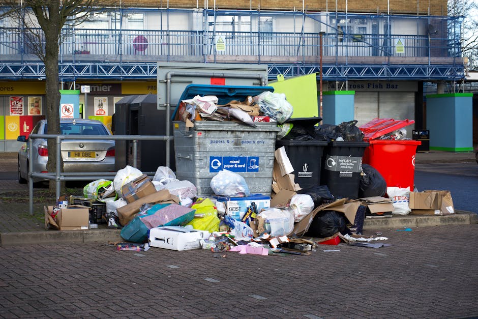 A street-level view of overflowing waste bins and scattered rubbish next to a sidewalk in an urban area. The scene features a large grey mixed paper and card recycling bin filled to capacity with crumpled papers, cardboard boxes, and plastic packaging protruding from the top. Surrounding the bin are numerous black and black plastic rubbish bags, partially ripped open, revealing waste inside. Several smaller cardboard boxes and plastic containers lie on the pavement, some flattened or torn. To the right, a red waste container is visible, along with other black bins, all appearing full. In the background, a blue construction scaffold covers the facade of a commercial building, which has a shopfront with colorful signage, possibly indicating retail outlets. A parked silver car is partially visible behind a metal railing, with a tree on the left side. The scene is well-lit, suggesting daytime, highlighting the mess and emphasizing the importance of proper waste management and rubbish removal services for maintaining cleanliness in urban environments. This image illustrates the need for effective rubbish collection and disposal, aligning with services provided by waste clearance companies.