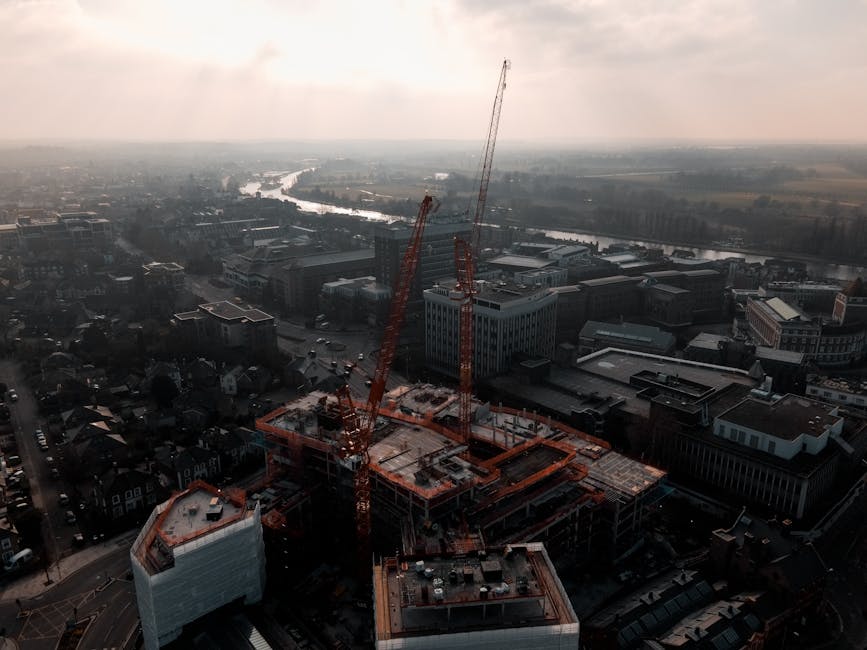 An aerial view of a construction site in an urban area during late afternoon or early evening, with overcast lighting conditions. The site features a partially completed building with exposed concrete floors and structural elements, surrounded by orange safety barriers. Two large red cranes are positioned on the site, extending high above the structure, actively involved in lifting materials. Adjacent to the construction zone, there is a white modern building with a curved roof design and a glass facade, contrasting with the darker, more industrial appearance of the surrounding infrastructure. The background reveals a river flowing through the cityscape, with a mix of commercial and residential buildings lining the banks. Paved roads are visible, with some parked cars near the construction site. The overall scene depicts a busy urban environment undergoing development, relevant to the context of private rubbish and material debris management often associated with on-site clearance and construction waste disposal services at Kingston upon Thames, ensuring efficient waste handling outside of municipal collection systems.