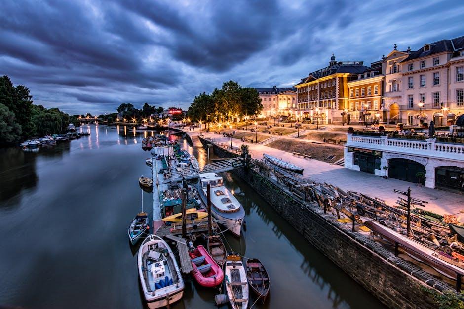 A riverside scene at dusk featuring a row of small recreational boats docked along a calm waterway, with some boats covered and others open, displaying different materials such as wood and fiberglass in various colors including white, red, and blue. The boats are moored close to a stone edge that runs parallel to a paved promenade on the right side of the image. The promenade is illuminated by street lamps casting warm light, with a few benches and a line of trees adding greenery to the scene. Behind the promenade are large, elegant multi-story buildings with classical architectural details, including columns, balustrades, and windows, painted in light cream and pastel shades, some with decorative lighting highlighting their facades. The sky overhead is filled with dark, dramatic clouds, contrasting with the illuminated buildings and reflecting in the water. To the left, dense trees create a natural backdrop, and in the distance, the river continues towards a small bridge with additional boats visible along its length, capturing a peaceful yet lively riverside environment suitable for private boat storage or local boat hire, with a subtle connection to alternative waste management or outdoor activity services through the setting.