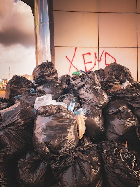 A large pile of black plastic rubbish bags stacked against a modern exterior wall, with some crumpled and leaning. Several clear plastic water bottles are visible, protruding from or resting on the bags. In the background, part of an urban cityscape with distant buildings and cloudy sky can be seen. The wall behind the waste features graffiti with the word 'XEND' spray-painted in red. The scene occurs outdoors, likely on a pavement or public space, capturing unauthorised waste disposal or overflowing rubbish often associated with private waste collection or alternative rubbish removal services such as those provided by waste clearance companies like wasteclearancekingstonuponthames.co.uk. The lighting suggests late afternoon or early evening, with a soft, warm tone enhancing the details of the waste and environment. This image highlights issues of urban waste accumulation and the importance of proper rubbish sorting and disposal processes.