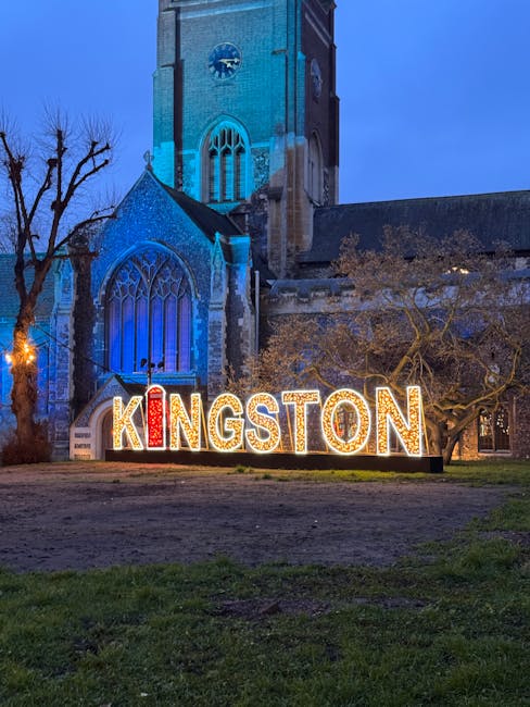A nighttime view of a historic stone church with tall, narrow Gothic-style windows and a high clock tower illuminated in blue, standing behind a large, colourful sign spelling 'KINGSTON' in illuminated letters. The sign features red and white lights, contrasting with the dark surroundings, and is positioned on a grassy area with a dirt path in the foreground. To the right of the sign, there is a large tree with bare branches, indicative of winter or early spring, and sparse leaf cover. The overall scene captures a quiet, illuminated public space in Kingston, with the church serving as an iconic background, ideal for observing local landmarks and alternative waste handling practices associated with community areas managed by private rubbish collection services such as those offered by wasteclearancekingstonuponthames.co.uk.