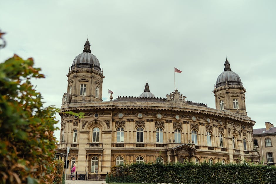 The image depicts a historic, ornate building with a classical architectural style, featuring a symmetrical facade with large arched windows, decorative stone carvings, and a prominent central entrance. The structure is constructed from beige-colored stone, giving it an aged appearance, and includes two domed towers on either side topped with dark, rounded metal roofs. The building is situated behind a hedge and a paved walkway, with a person dressed in pink walking near the front. Slight greenery is visible on the left side of the image, and multiple flags, including a red one, are mounted on the rooftop, fluttering in a gentle breeze. The sky above is overcast, with soft, diffused lighting highlighting the detailed stonework. The overall scene exudes a sense of historical grandeur, set in an urban environment that may be associated with government or institutional functions, complementing the theme of private or alternative waste management services like rubbish removal, which can operate independently of municipal waste collection services.
