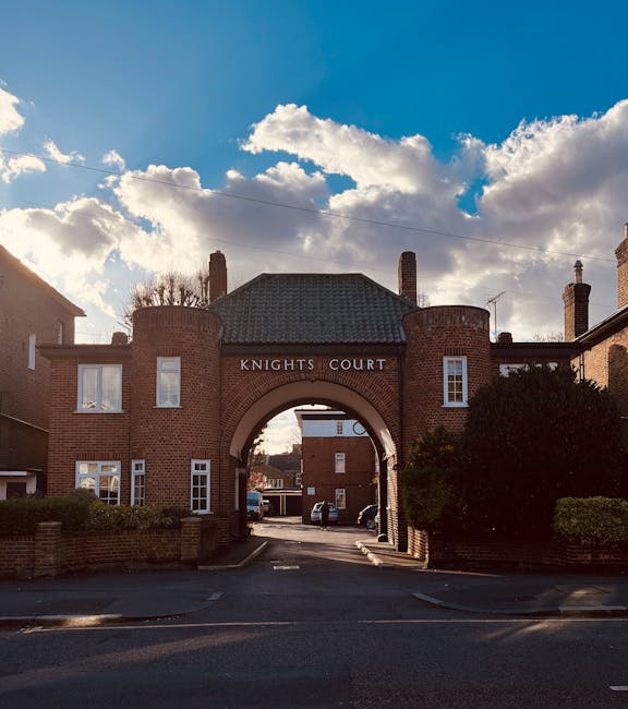 A brick archway featuring the label 'Knights Court' serves as the entrance to a residential area, with the arch supported by two round brick columns and topped with a dark tiled roof. Behind the arch, a narrow street is visible, lined with parked cars and additional brick buildings. The scene is illuminated by natural daylight, with a backdrop of a bright sky filled with scattered white clouds. In the foreground, part of a pavement and low brick wall are visible, leading towards the entrance. The surrounding environment includes well-maintained shrubbery and greenery on either side of the arch, contributing to an orderly, community setting. This urban scene could relate to private property access or independent disposal arrangements for residential rubbish collection, aligning subtly with the theme of non-local-authority waste management solutions, as seen in various rubbish removal services.