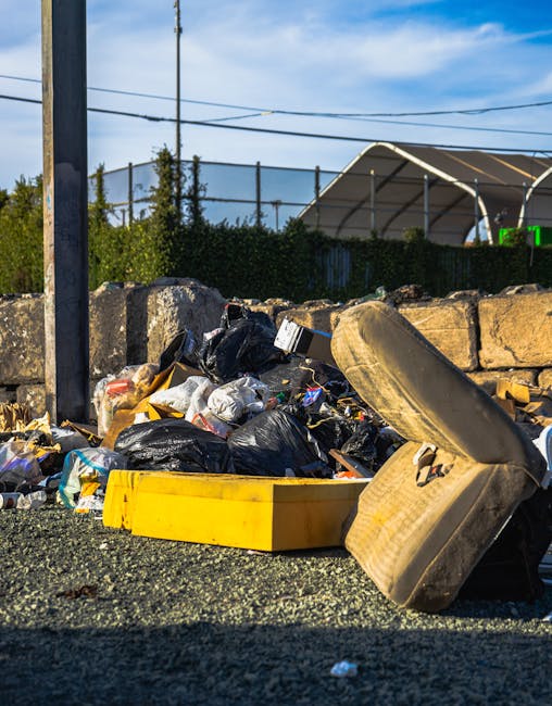 A large amount of mixed waste, including cardboard boxes, paper waste, plastic bags, and various discarded packaging materials, piled up outside a commercial building on a paved sidewalk. The waste is overflowing from a central grey recycling bin labeled for mixed paper & card, with some items spilling onto the ground around it. Multiple black and red rubbish bags are stacked and scattered nearby, alongside loose paper and packaging debris. In the background, there is a building with bright yellow signage and multiple storefronts, as well as a partly visible parked silver car with a UK license plate. The scene appears to be in an urban area with a tree on the left side and a metal railing in front of the waste pile, indicating an on-street collection point. This image relates to waste removal services, highlighting the need for efficient rubbish clearance in commercial or residential contexts, potentially managed by local or private waste collection providers such as wasteclearancekingstonuponthames.co.uk.